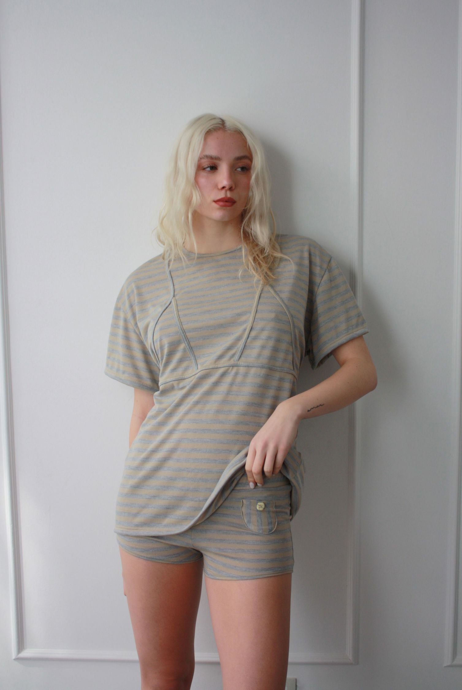 Woman stands against a wall lifting up one end of an oversized fitted cotton tee. The tee is cinched at the waist with a bikini detailing and babydoll top silhouette.